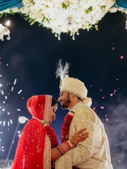 The couple shares a look of love under a grand floral chandelier, with sparks flying around them. This image combines intimacy with the spectacle of a grand Indian wedding.