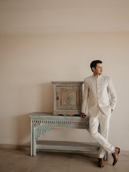 A stylish solo portrait of the groom in an elegant white bandhgala suit. The simple background and antique furniture add a touch of classic sophistication.