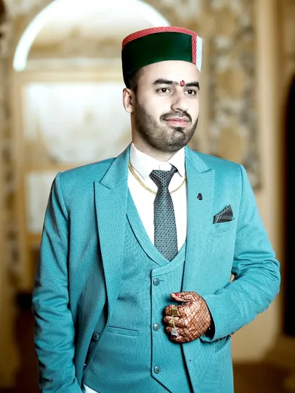 A groom looking sharp in his three-piece suit and traditional Himachali topi. The warm lighting highlights the texture of the suit and the intricate henna on his hand.