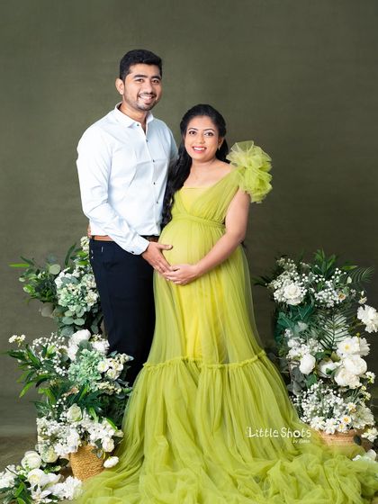 A lovely couple's portrait featuring a floral arrangement. The mom-to-be is in a beautiful lime green gown, standing with her partner in a studio setting adorned with white flowers.