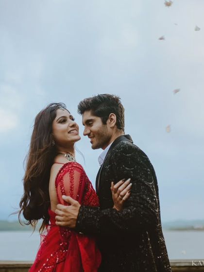 A candid moment of connection as petals fall around the couple during their lakeside pre-wedding session. The woman's vibrant red gown stands out beautifully against the soft sky.