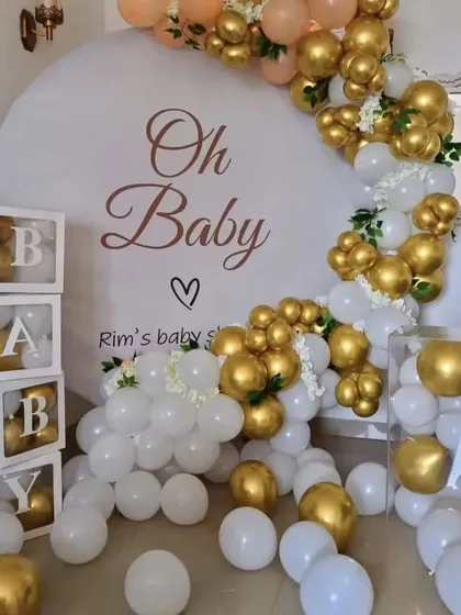 An elegant baby shower setup with a white and gold theme. The large backdrop is personalized for the mom-to-be, and the decor includes "BABY" blocks and a beautiful balloon arrangement.