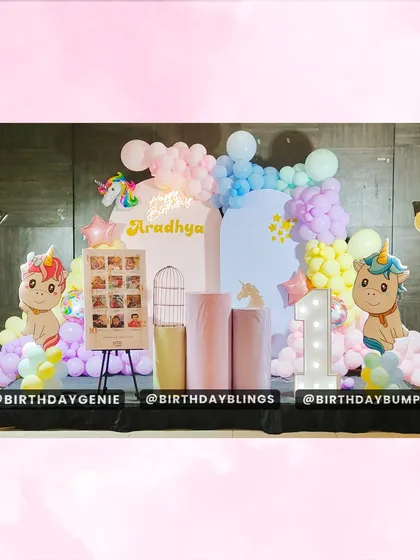 A sweet and colorful unicorn-themed first birthday party. The stage is decorated with pastel rainbow balloons, cute unicorn cutouts, and a light-up number '1'.