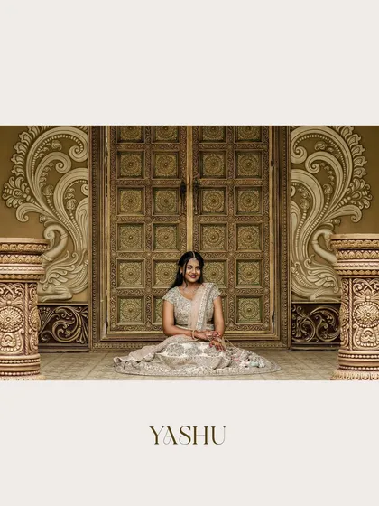 A stunning portrait of a bride-to-be seated before ornate, traditional doors. Her sparkling lehenga and happy smile make this a grand and joyful engagement photo.
