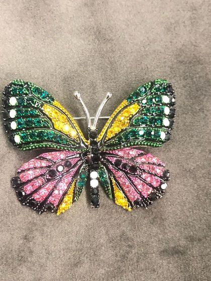 A beautiful butterfly brooch, intricately detailed with green, yellow, and pink stones. Accessories like this add a complete and stylish look to any outfit, from a simple scarf to a formal blazer.