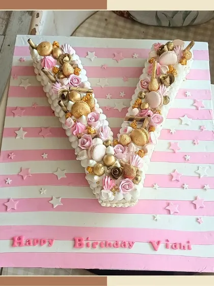 A creative and beautiful monogram cake in the shape of the letter 'V', decorated with macarons, rosettes, and golden pearls.
