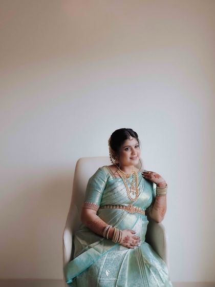 A graceful indoor portrait of a mother-to-be in a light blue silk saree. The clean, minimalist background emphasizes her elegance and the beauty of her traditional attire.