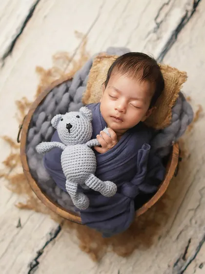 This little one is dreaming soundly in a rustic wooden bowl, cuddling a small knitted teddy bear. The textures of the wood, wool, and wrap create a cozy, tactile image.