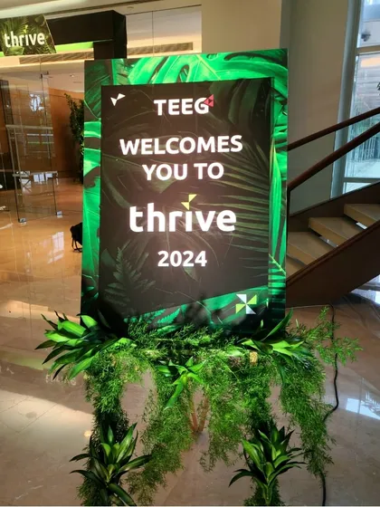 A branded welcome sign for the 'thrive' corporate summit. The sign is designed as a digital screen, framed by lush tropical foliage that ties in with the event's visual theme.