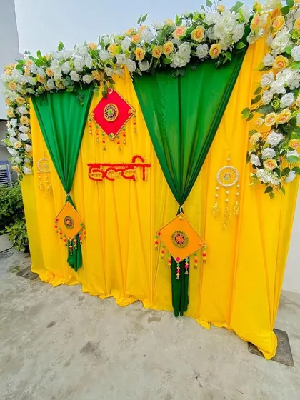 A classic Haldi backdrop I set up outdoors, using vibrant yellow and green fabrics. The decor is accented with white and yellow floral arrangements on top and traditional hangings, with a 'Haldi' sign in the center.