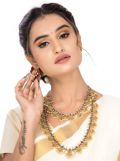 This close-up focuses on the model adjusting her traditional earring, drawing attention to the detailed craftsmanship of the South Indian jewellery set. The white background ensures the gold and gemstones are the hero of the image.
