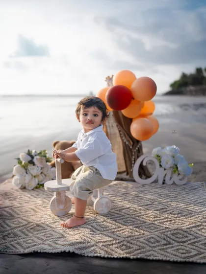 Celebrating a first birthday with a boho-themed setup on the beach. This sitter session captures the baby's curious spirit with adorable props like a wooden tricycle, balloons, and a 'One' sign against the ocean backdrop.