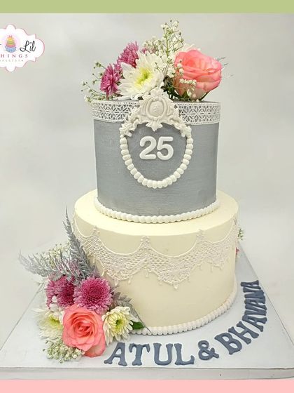 A grand 25th anniversary cake for Atul and Bhavna, featuring silver and white tiers with intricate lace details and fresh flowers.