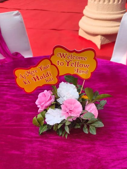 A fun and quirky centerpiece for a Haldi party table, with a small floral arrangement and signs that say "Aaj Mere Yaar ki Haldi hai".