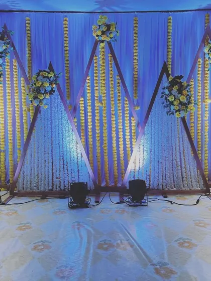 A modern and geometric stage design for a Sangeet or reception. The backdrop features triangular frames decorated with white and yellow flower strings.