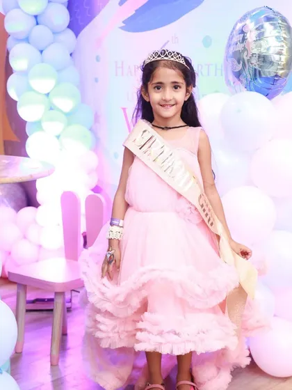 The birthday princess in her element. Dressed in a beautiful pink gown and a "Birthday Girl" sash, she's ready to celebrate in front of a mermaid-themed balloon backdrop.