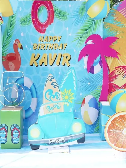 A vibrant, tropical-themed photo backdrop for a 5th birthday pool party. The display features a vintage car cutout, surfboards, and a wall of fresh coconuts for guests.