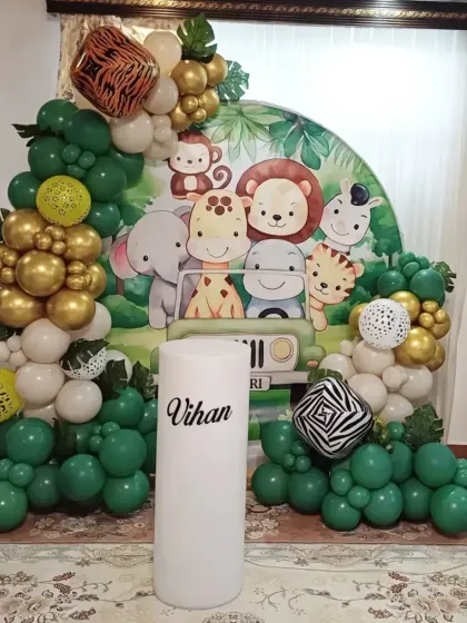 A cute safari-themed decoration for a second birthday. This setup includes a backdrop with cartoon jungle animals, balloon arrangements in green and gold, and personalized name blocks.