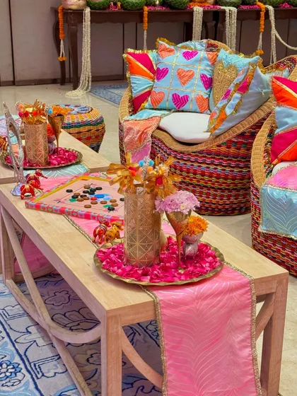 A cozy seating corner from our Garba celebration decor. The setup includes wicker chairs with colorful, heart-embroidered cushions, a wooden coffee table with a pink runner, and trays of flower petals.