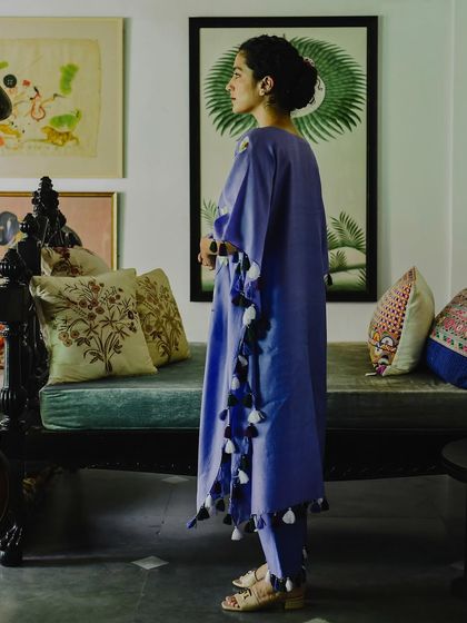 A full-length side profile of a model wearing a lavender kaftan set from Baavli. The shot is set in a beautifully decorated room, showing how the garment moves and drapes in a real, lived-in space.