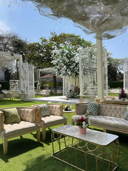 An elegant outdoor lounge area with a custom-built white floral tree as the centerpiece. The design combines comfortable seating with artistic floral installations for a relaxed yet sophisticated reception.