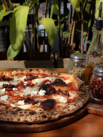 Our Smokey Marzano pizza served outdoors, accompanied by classic condiments like chili flakes and oregano. The natural light highlights the fresh feta and sundried tomatoes.
