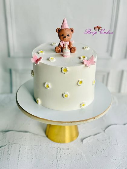 A sweet and delicate cake featuring a tiny teddy bear having a party. The cake is decorated with small daisies and butterflies, perfect for a simple, heartfelt celebration.