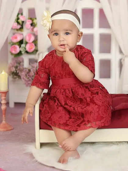 A toddler girl in a beautiful red lace dress sits on a miniature bed in a romantic, floral-themed room.