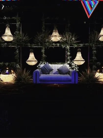 A moody and romantic stage setup for an engagement, featuring a simple blue sofa surrounded by greenery and hanging crystal chandeliers against a dark backdrop.
