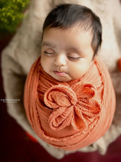 A close-up of a newborn wrapped in a warm orange swaddle, looking like a tiny, precious flower bud. The details of the wrap and the baby's peaceful face are the focus.