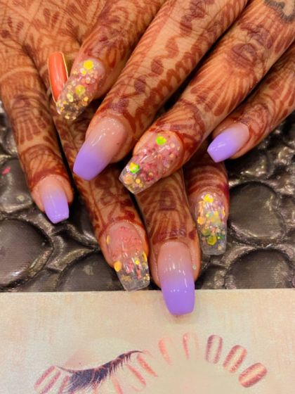 A festive and colorful design on henna-adorned hands. The nails feature a purple ombre, with accent nails covered in chunky, multi-colored glitter.