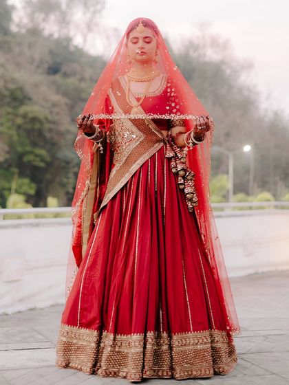 A full-length portrait of the bride holding her veil. The simplicity of the pose highlights the intricate details of her red and gold lehenga.