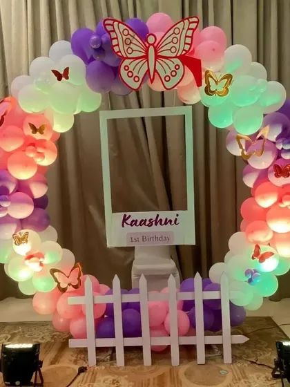 A butterfly-themed first birthday photo booth with a circular balloon frame and a polaroid-style cutout.
