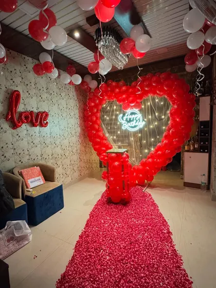 For romantic surprises, we can transform any room into a beautiful celebratory space. This setup features a large heart-shaped balloon arch, a 'Happy Birthday' neon sign, and a path of rose petals for an unforgettable surprise.