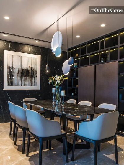In this dining room, a dark marble table is surrounded by soft, modern chairs, creating a dramatic yet inviting atmosphere. The space is defined by a moody color palette, abstract art, and sculptural pendant lights that add a touch of elegance.