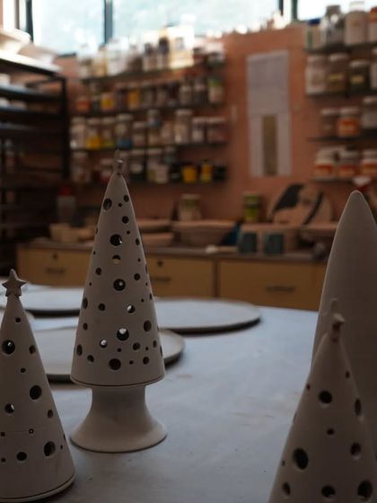 Unfired ceramic Christmas trees with cutwork details, sitting on a studio table waiting for the kiln. The background shows our shelves stocked with glazes and tools.