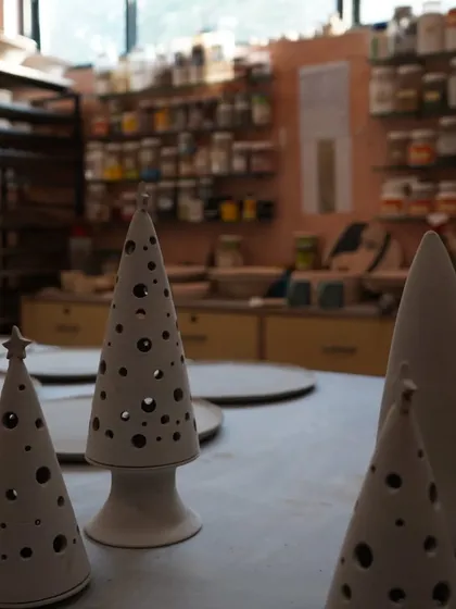 Unfired ceramic Christmas trees with cutwork details, sitting on a studio table waiting for the kiln. The background shows our shelves stocked with glazes and tools.