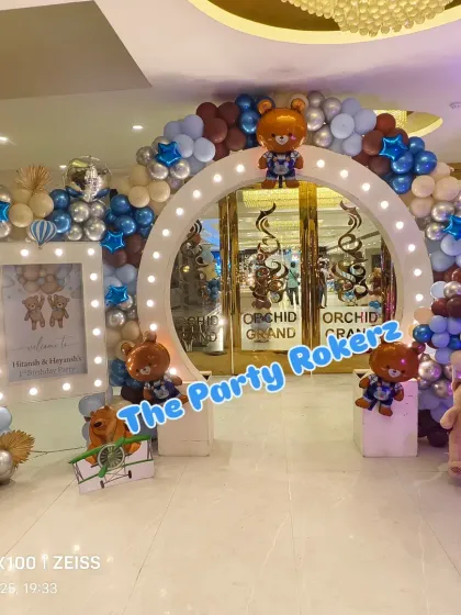 A grand entrance for a Teddy Bear-themed first birthday. The lighted arch is decorated with brown, blue, and beige balloons and cute teddy bear cutouts.