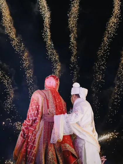 A dramatic shot from behind the couple during their varmala, with fireworks creating a stunning, fiery halo around them.