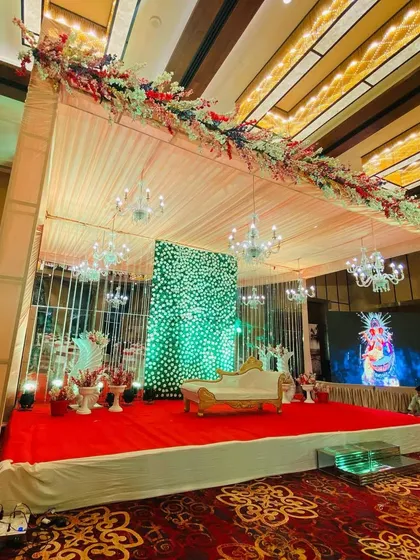 A grand indoor stage setup in a banquet hall, featuring a large green floral wall and elegant chandeliers.