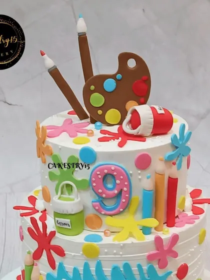 A creative two-tier artist-themed cake for a 9th birthday. This 3kg blueberry cake is decorated with fondant color splashes, paintbrushes, and a color palette.