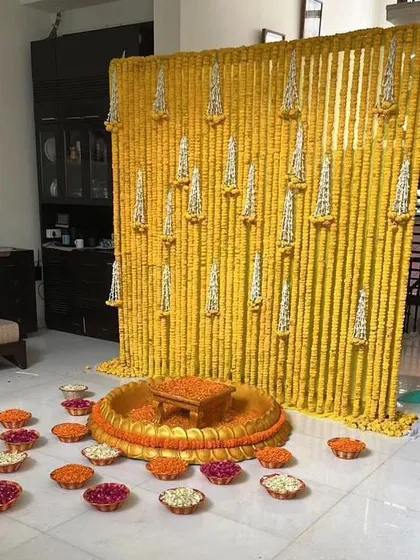 A simple yet elegant Haldi backdrop for an intimate home ceremony. The wall of marigold strings is accented with hanging tuberose tassels, and the floor is decorated with small bowls of flowers.