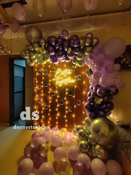 We designed this romantic proposal setup featuring a 'Better Together' neon sign. The backdrop includes a curtain of fairy lights with polaroid-style photos clipped on, framed by a lush balloon arch in shades of purple and chrome.