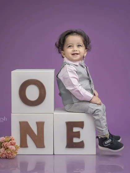 A dapper little gentleman celebrating his first birthday. He's sitting on 'ONE' blocks in a smart vest and shirt, giving a charming smile for his milestone portrait.