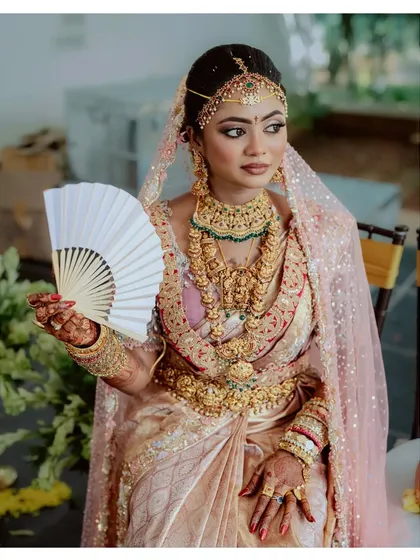 A bride in a stunning pastel saree, holding a fan, looks every bit a queen on her special day.