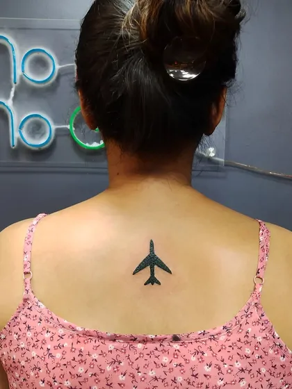 A small, solid black airplane tattoo on the upper back. A perfect minimalist design for a travel lover or someone in aviation.