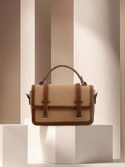 A tan canvas and leather bag set against a background of neutral-toned vertical stripes. The lighting and composition are warm and sophisticated.