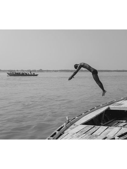 A black and white photo of a man diving headfirst from a boat into the Ganga. The composition captures the peak of his dive, with another boat in the distant background.