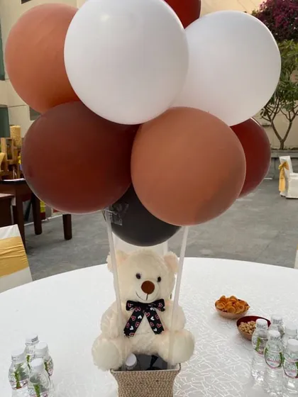A charming table centerpiece featuring a teddy bear in a hot air balloon basket, using earthy tones of brown, white, and black balloons.