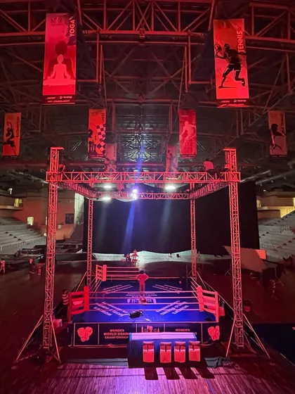 A wide shot of the boxing ring setup under the stadium's high ceiling. The hanging banners and red lighting create a powerful visual, showcasing the grandeur of the event.